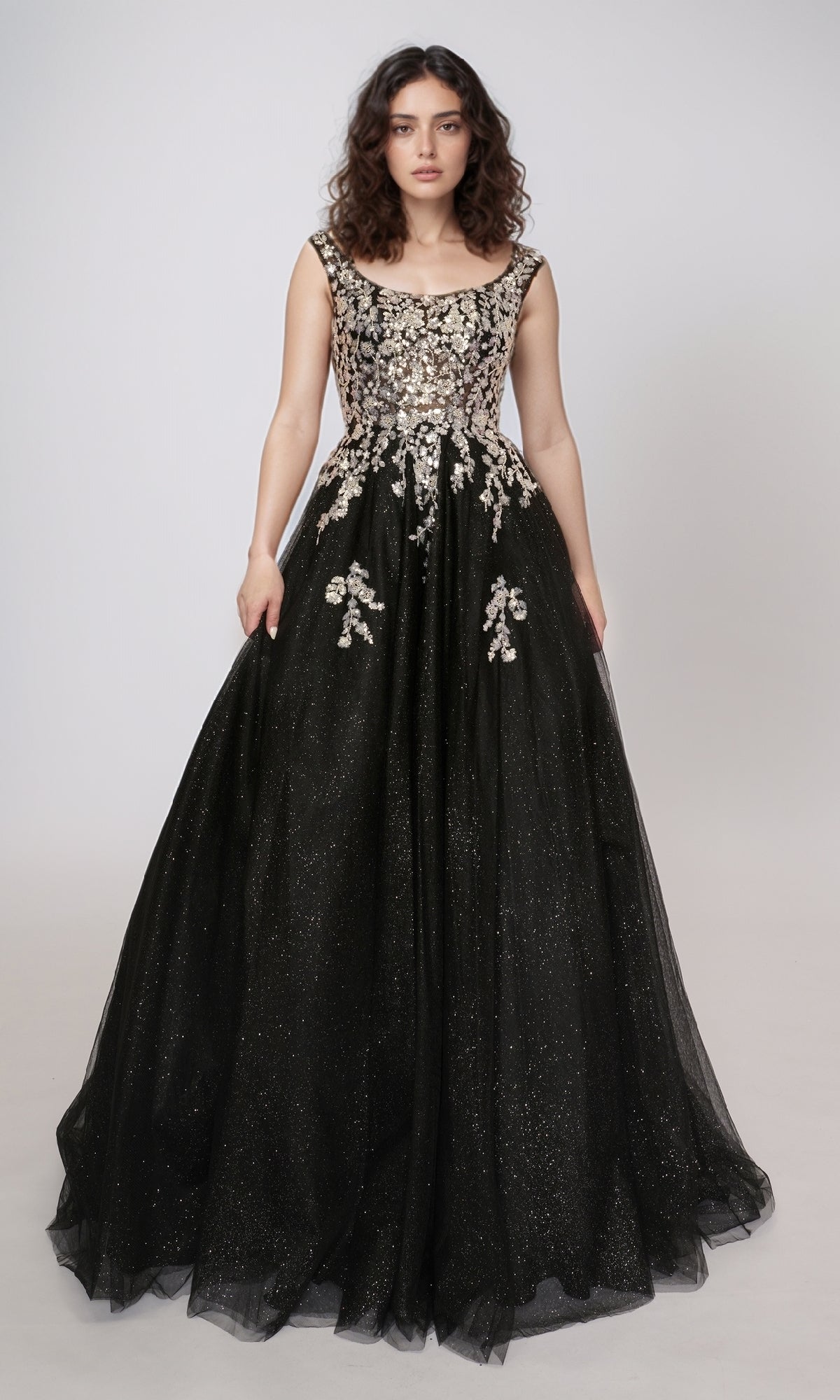 A woman in the Nina Canacci 3266 long prom dress—black glitter tulle with a fitted, embellished bodice and floral appliqués—stands against a plain light background.